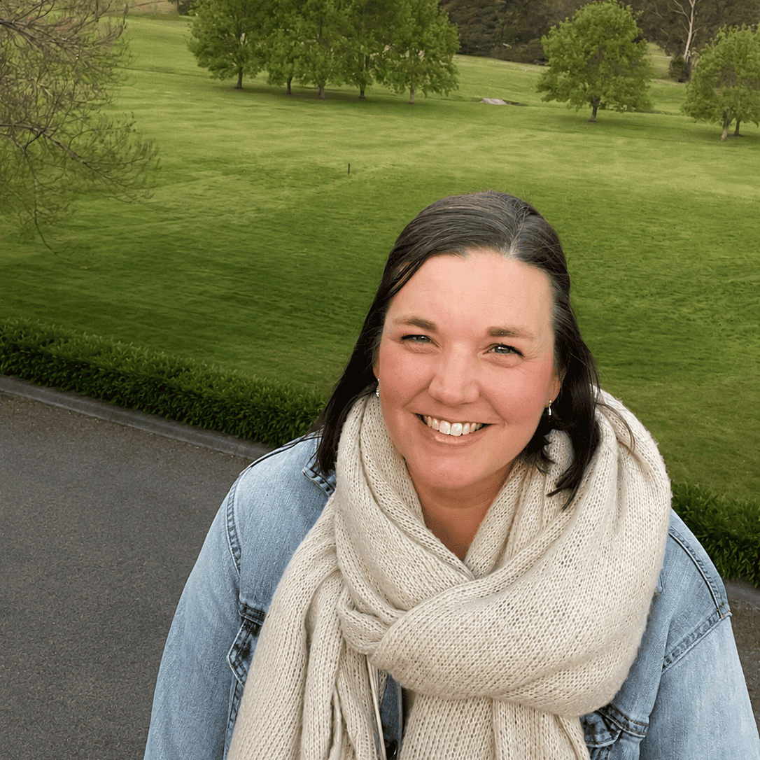 A smiling woman outdoors in front of a green park landscape, wearing a light beige knit scarf and a blue denim jacket.